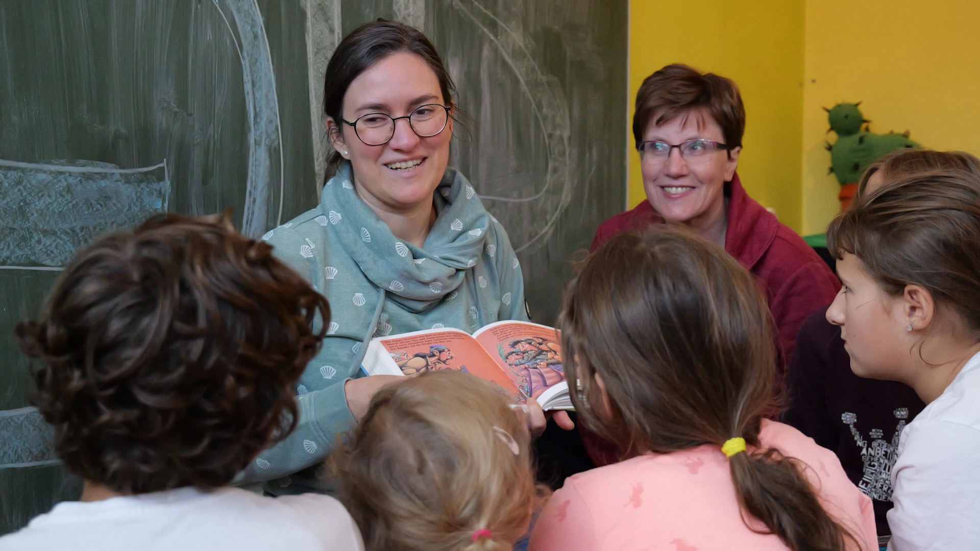 Person hält guckend vor mehreren Kindern in einem Kinderraum ein Kinderbuch in der Hand