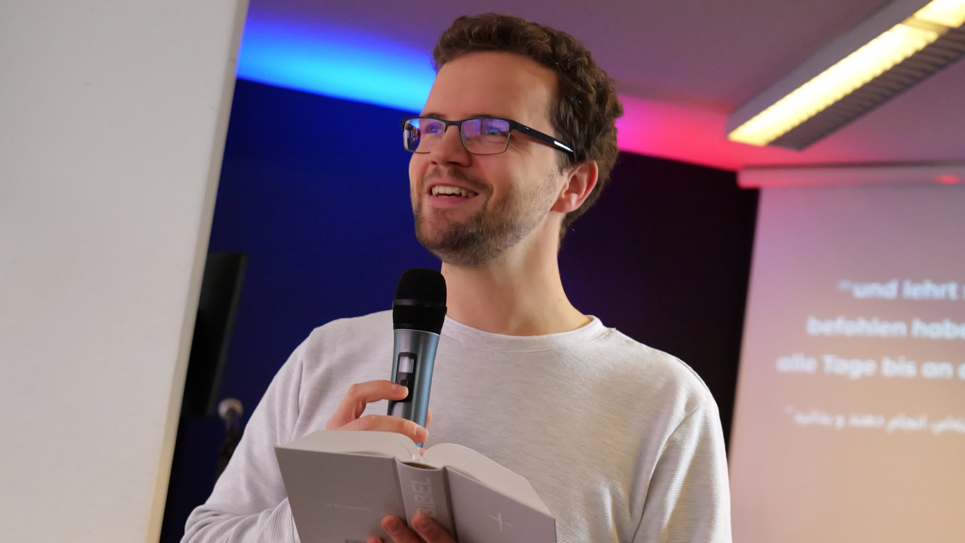 Benjamin Weis, Pastor der Elim Eichsfeld Gemeinde, hält ein Mikrofon und eine Bibel in der Hand und schaut zur Gemeinde lächelnd